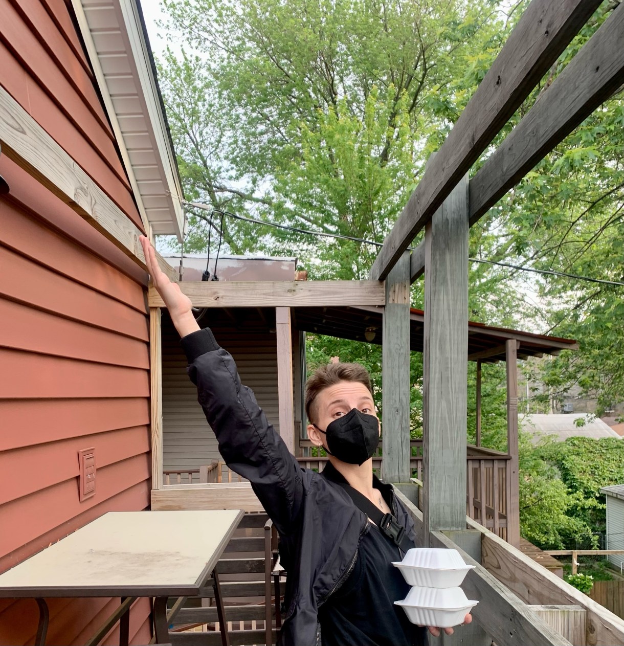 Ro gestures euphorically while holding what looks like some takeout containers. Ro is a white human with short brown hai wearing al black with an all black mask.