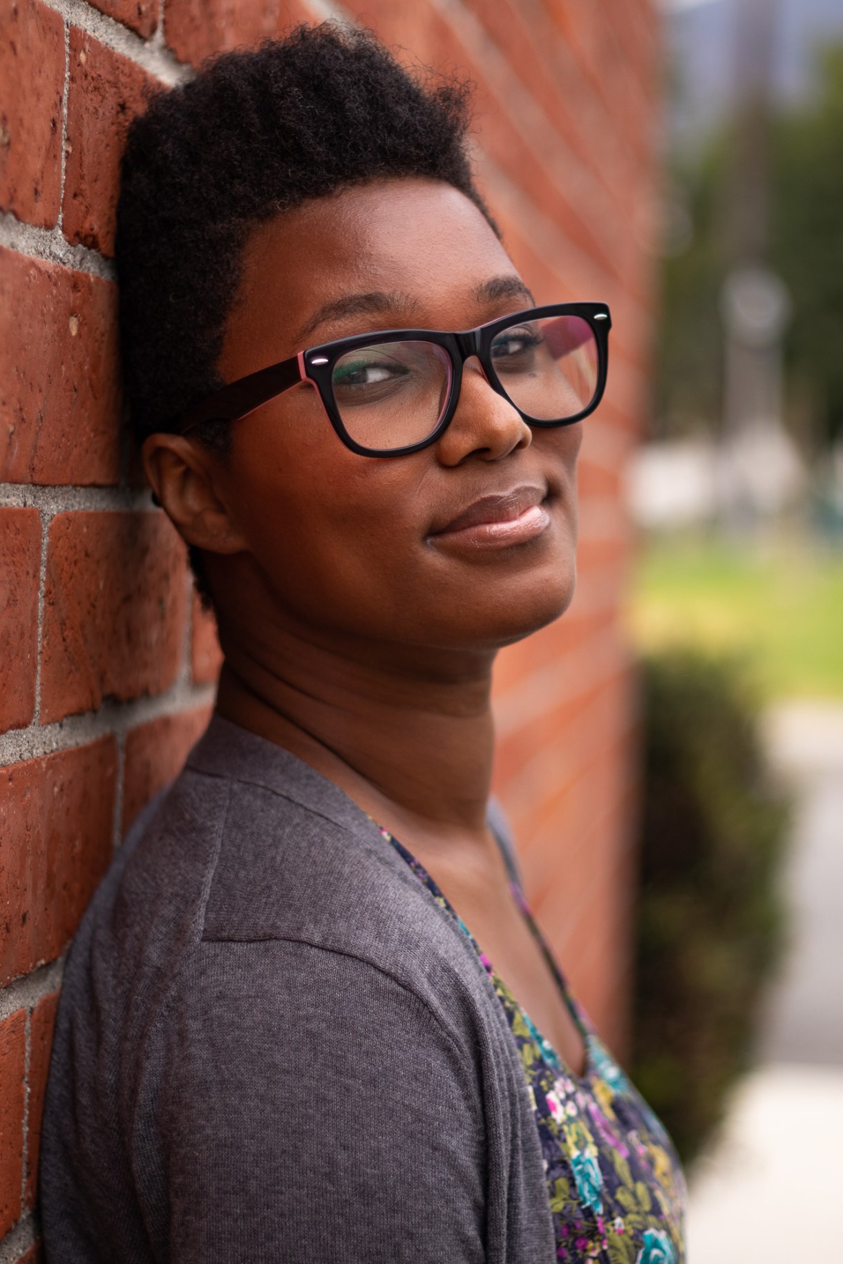 Sai, a Black woman with short black hair wearing a sweater and floral top and glasses, leans against a brick wall and smiles winningly