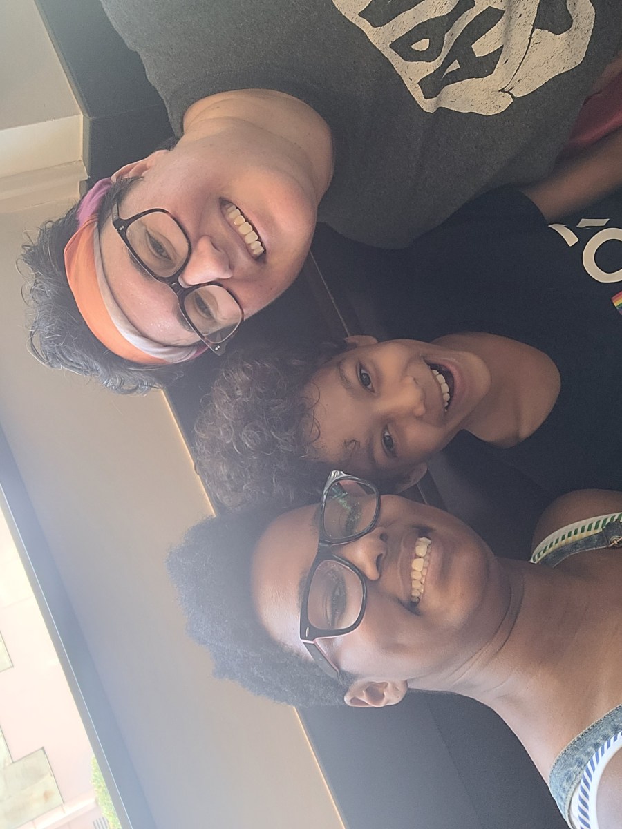 Sai, her fiancee Beth and her kid. Sai is a Black woman with short hair and glasses wearing a tank top. Beth is a white woman with short brown hair and glasses.
