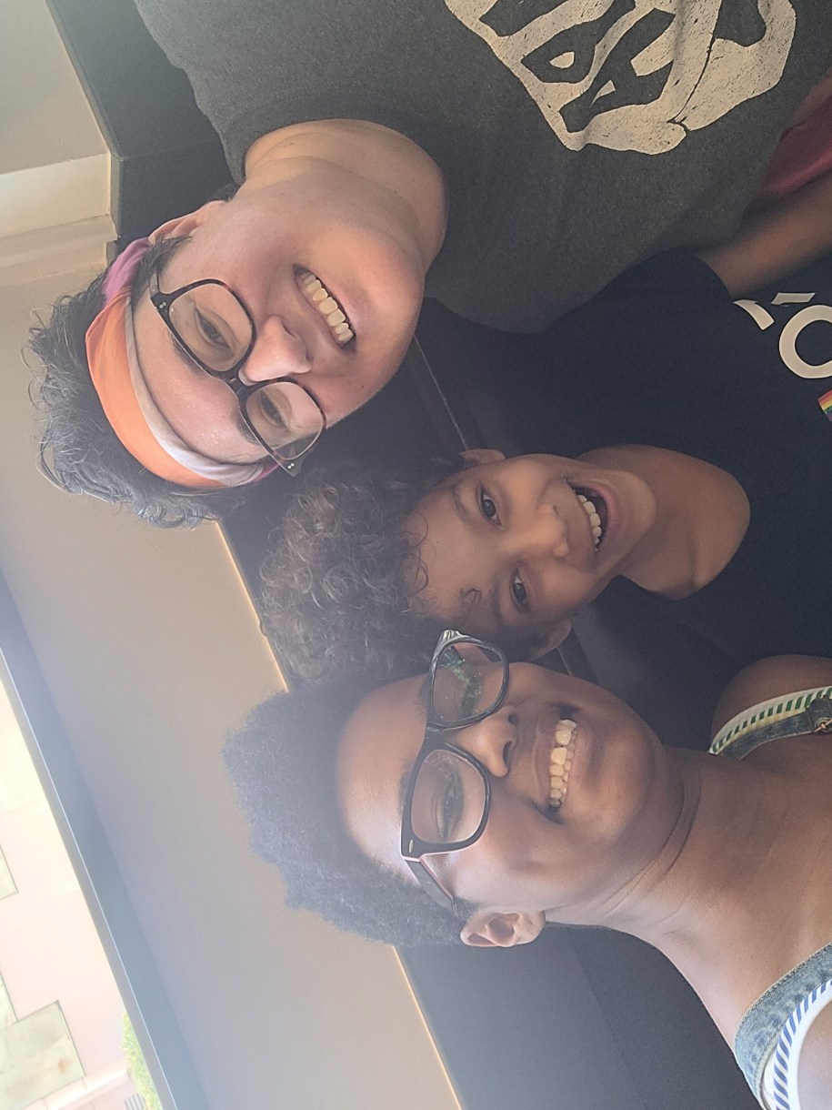 Sai, her fiancee Beth and her kid. Sai is a Black woman with short hair and glasses wearing a tank top. Beth is a white woman with short brown hair and glasses.