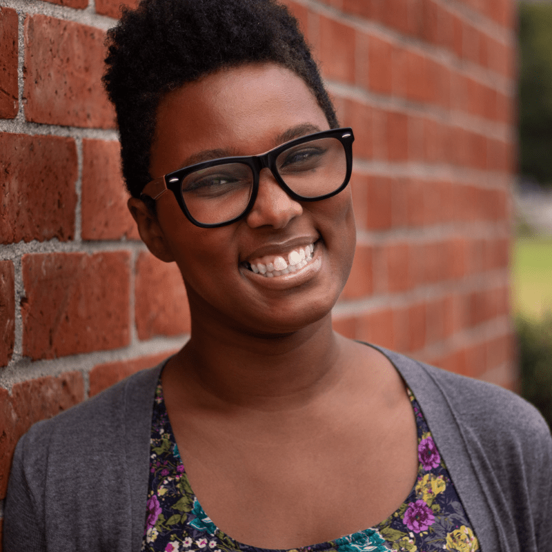Sa’iyda is a Black person with a high top fade and glasses, a grey sweater, smiling in front of a brick wall