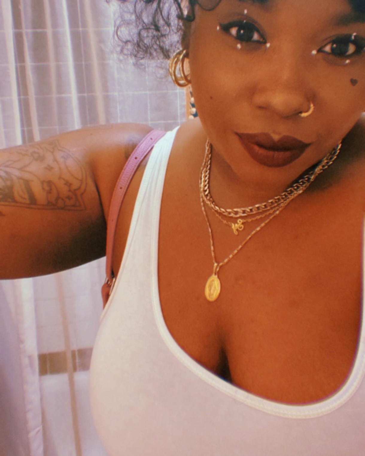 Shelli is wearing gold jewelr, a white tank top, and an amazing makeup situation with burgundy lips and white dots around her eyes. Shelli is a Black woman and a tattoo is visible on her arm.