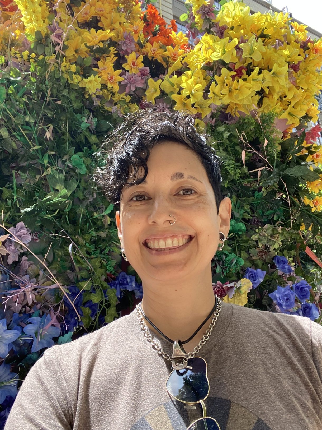 Tracy is here against a background of flowers. Tracy is an asian woman with curly dark hair, wearing a few necklaces and a tee shirt with multiple earrings, a big smile and a slit in her eyebrow.