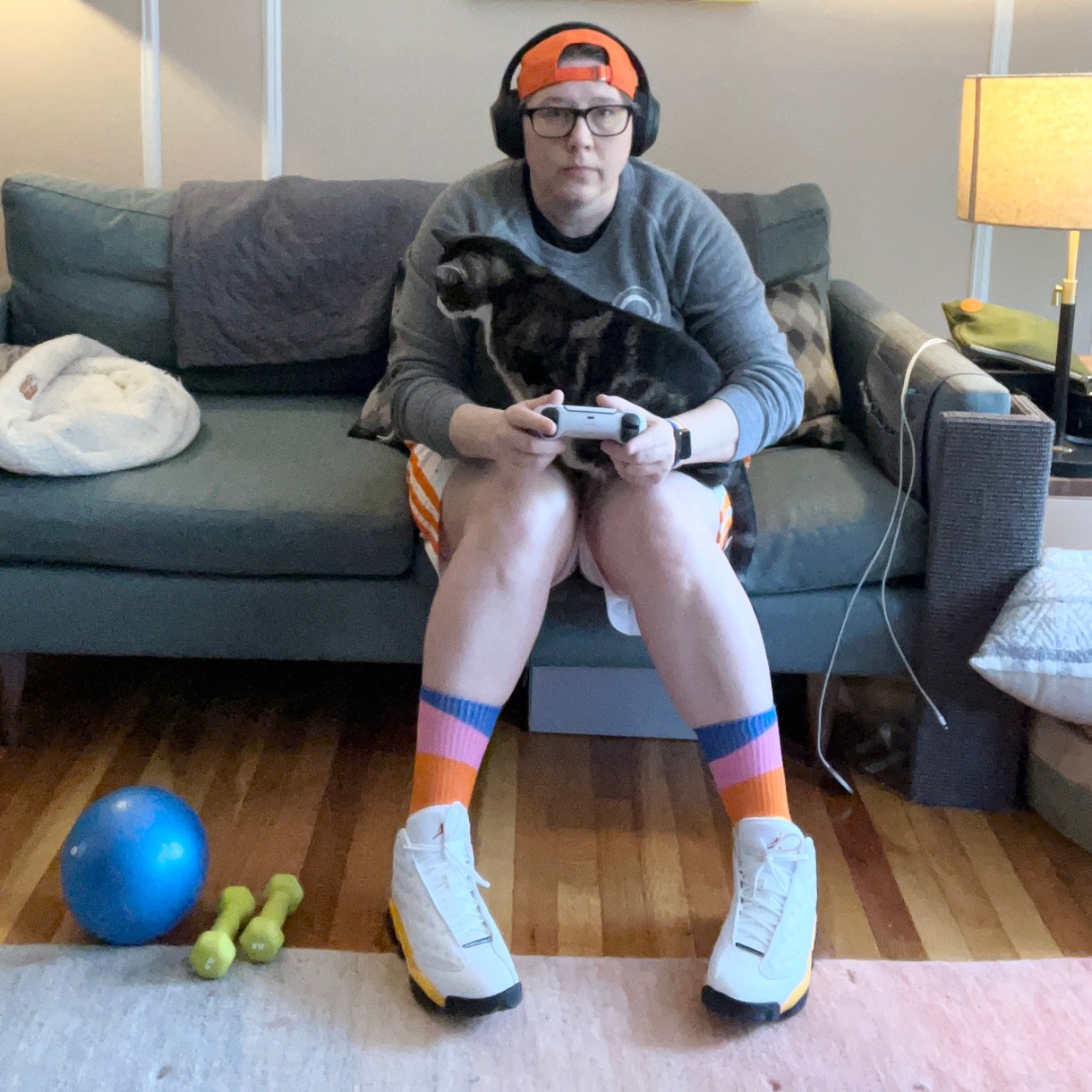 Heather is sitting her on her courch with one of her cats in her lap while playing video games. Heather is a white woman wearing a backwards orange baseball cap, headphones, colorful socks, basketball shorts and sneakers. Her cat is nuzzling her arm.