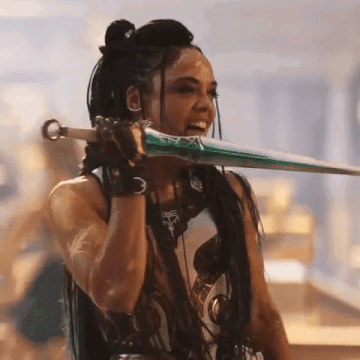 Tessa Thompson dressed as Valkyrie in "Thor: Love and Thunder," licking a sword with tongue during battle. Behind her is blurry gold buildings.