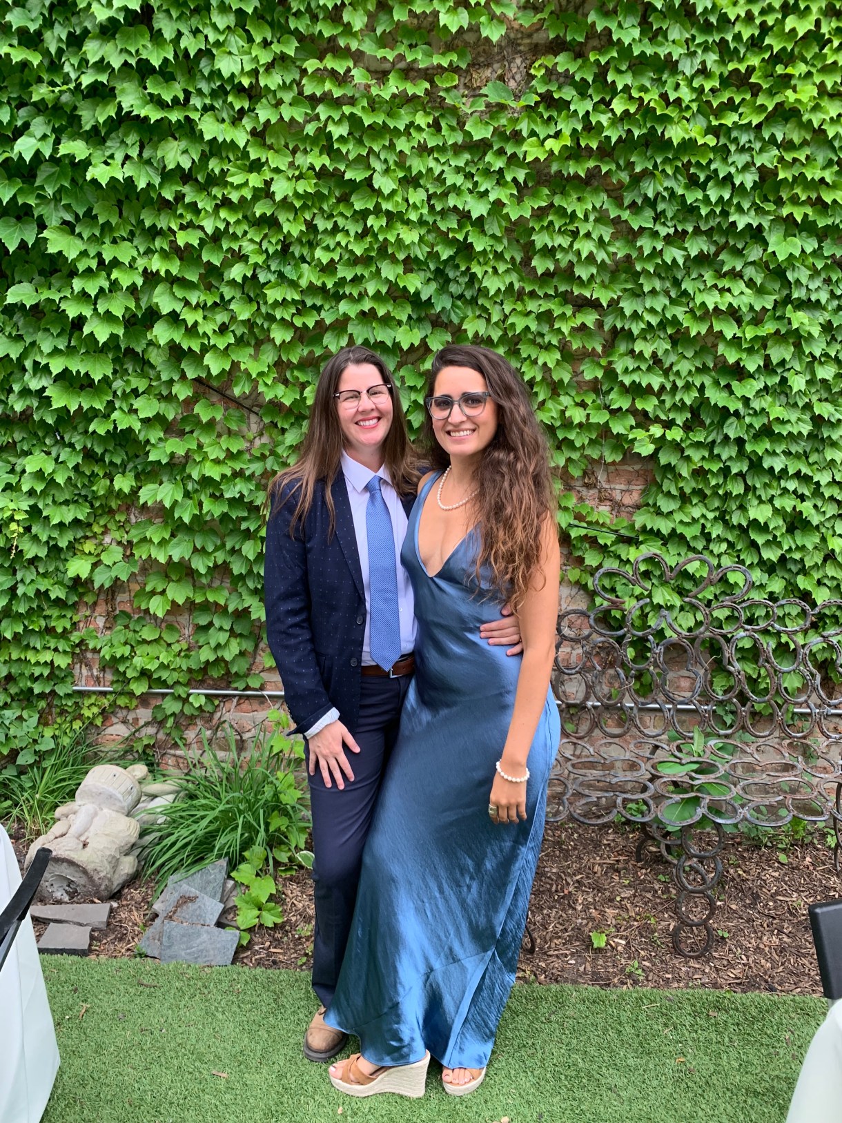 Kayla stands with her girlfriend Kristen in front of an ivy covered wall. Kristen is a white butch human wearing a blue suit. Kayla is a South Asian woman wearing a satiny blue dress with long brown hair and glasses.