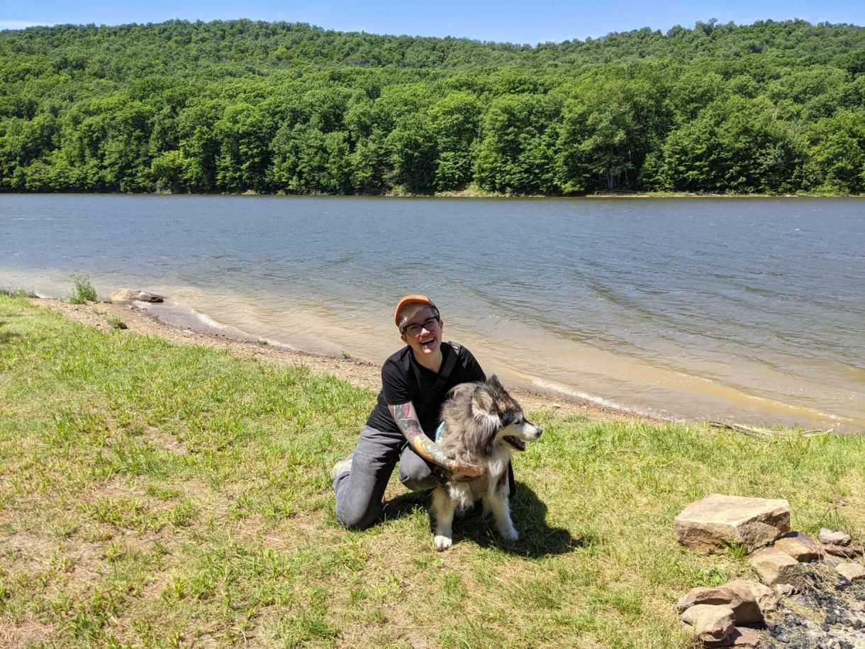 Nicole, a genderqueer white human kneels and holds their malamute mix rescue dog in front of a beautiful rippling blue river with a view of green trees on the other side. They are wearing a black tee shirt and gray jeans. Mya is trying to escape to jump in. Nicole has a tattoo sleeve visible on one of their arms.