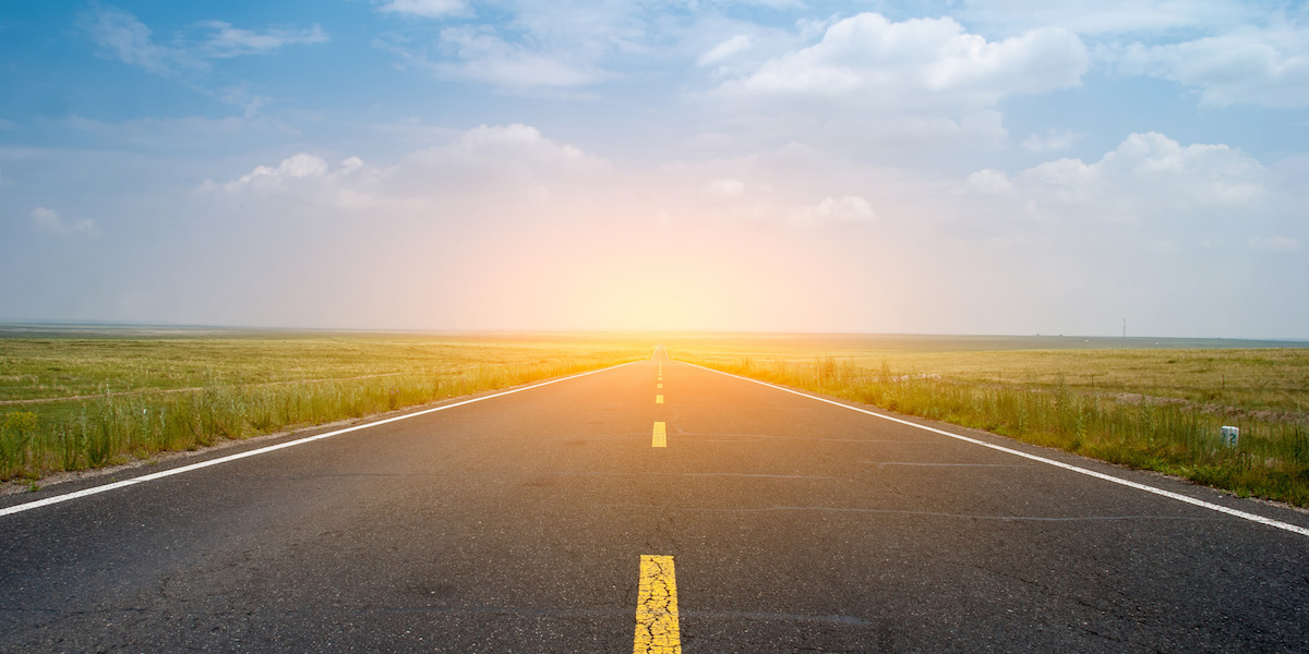 beautiful open road with green fields on either side, a blue sky, and the sun setting in the distance