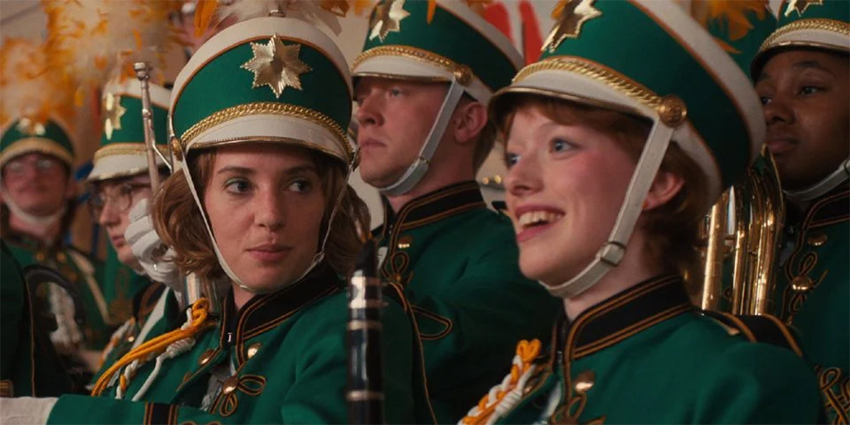 In a still from Stranger Things, Robin and Vickie are sitting side by side in their 1980s green marching band uniforms. Vickie is smiling off to the left of cameras and Robin is staring upon the side profile of Vickie's face intensely.