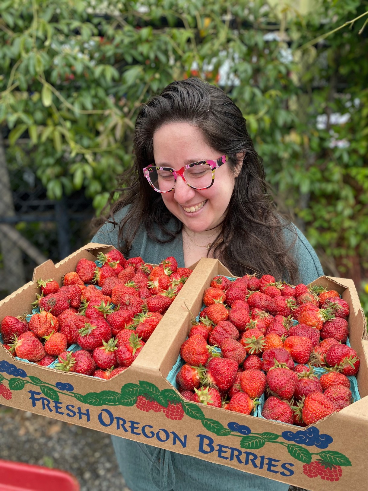 Vanessa is smiling down at a box of strawberries. She is a white woman with long brown hair and colorful floral glasses.