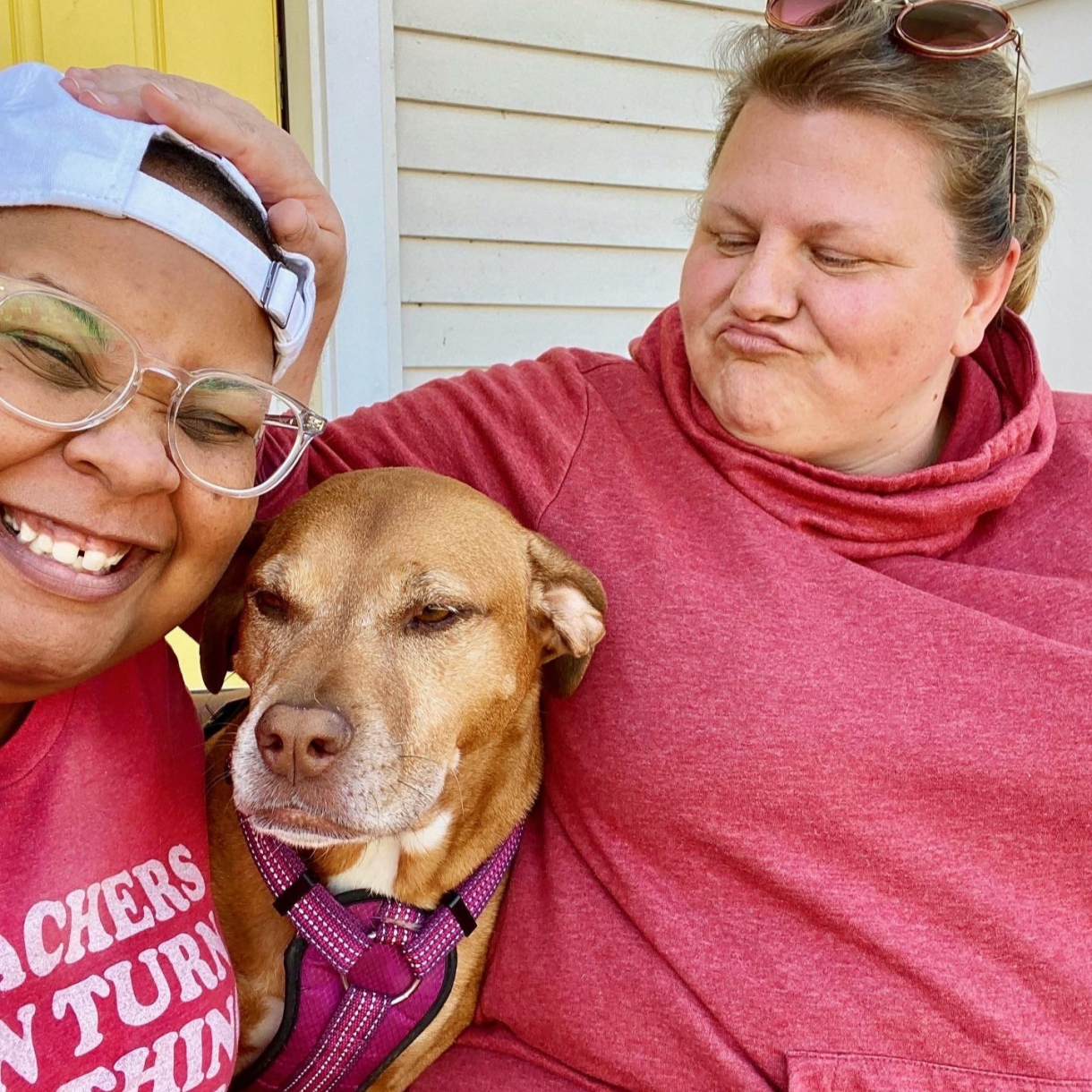 shea, a Black nonbinary human, and Jane, a white woman, pose with the dog, LilyPad. LilyPad has a tan coat. Both shea and Jane are wearing pink. shea also has on clear glasses and a backwards baseball cap. shea is smiling. Jane has her hand on shea's head and is kind of scrunching up her lips.