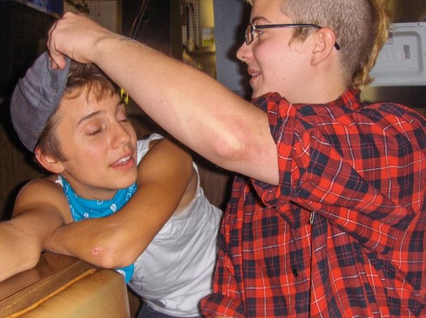 Ro (left), who has short brown hair, leans on a diner booth. They're wearing a white tank top and a bright blue bandana around their neck. Their eyes are closed. Eliot (right), who bleach blonde that's shaved on the side, wears glasses and a red flannel shirt. She reach over to Ro to remove Ro's grey newsboy cap.