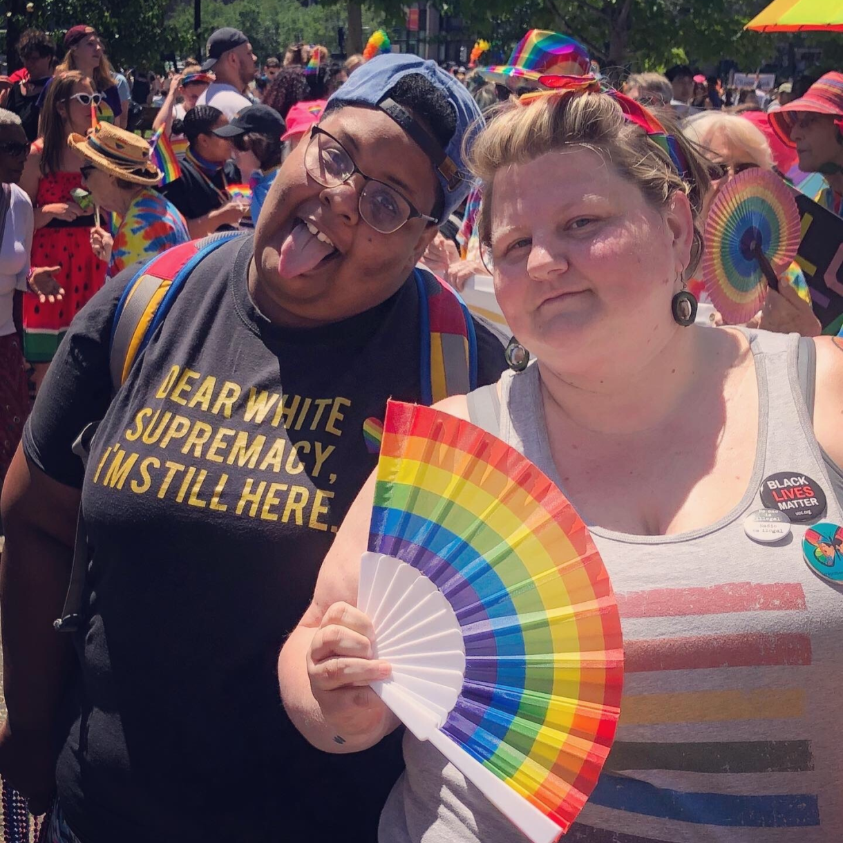 shea and Jane at a pride event. shea is wearing glasses, a backwards baseball cap, and a shirt that says "dear white supremacy, I'm still here" and Jane is wearing a shirt with several buttons including a "Black Lives Matter" button and is holding a rainbow fan and wearing a rainbow headband. a crowd of rainbow-clad people can be seen enjoying themselves behind them.
