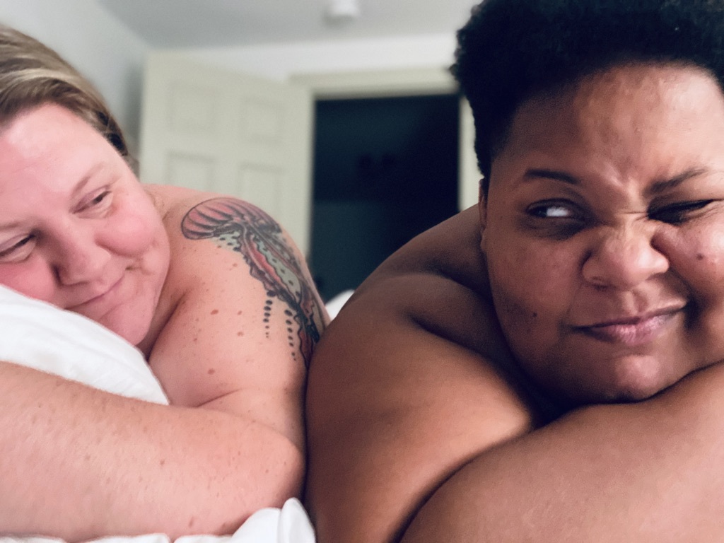 Jane and shea lie in bed. Jane smiles softly at shea. shea winks at Jane. They both aren't wearing any clothes! Jane is a white woman with blonde hair. shea is a Black nonbinary person with short black hari.