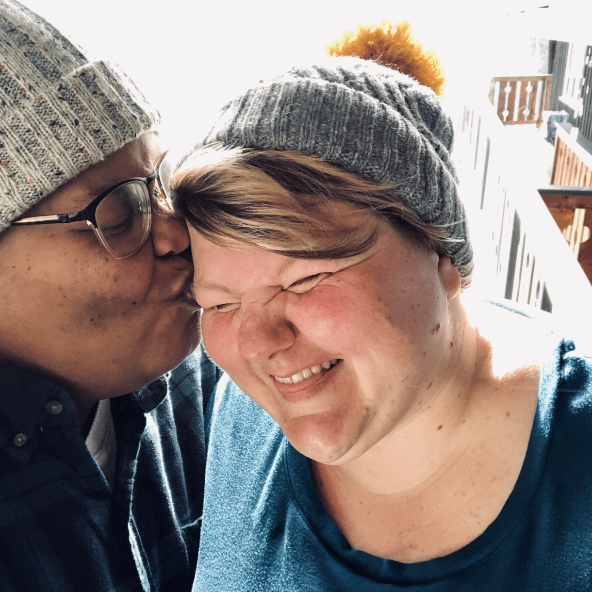 a close up photo of both shea and Jane in knit hats, smiling. shea kisses Jane on the cheek. 