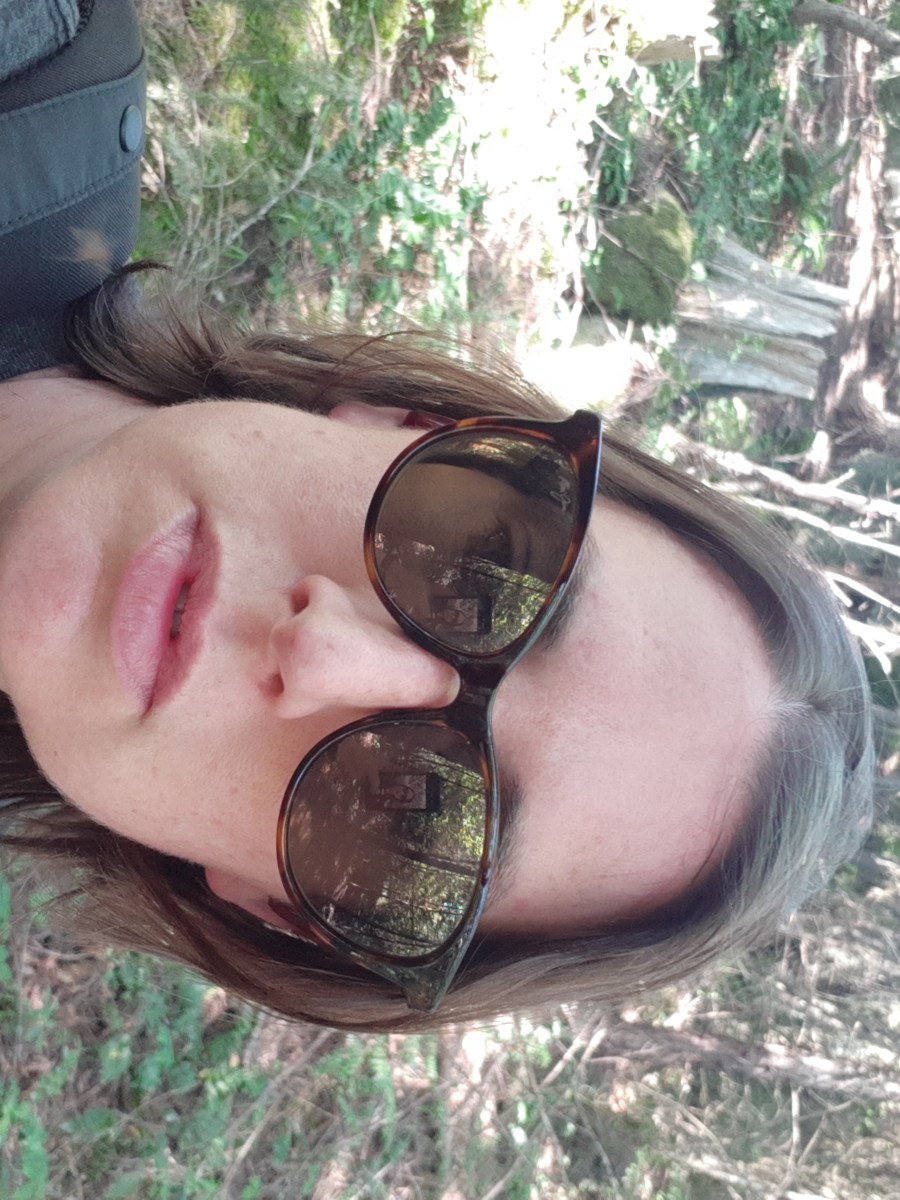 Casey, a white woman, regards the camera with lips parted. She is wearing sunglasses and has short brown hair.