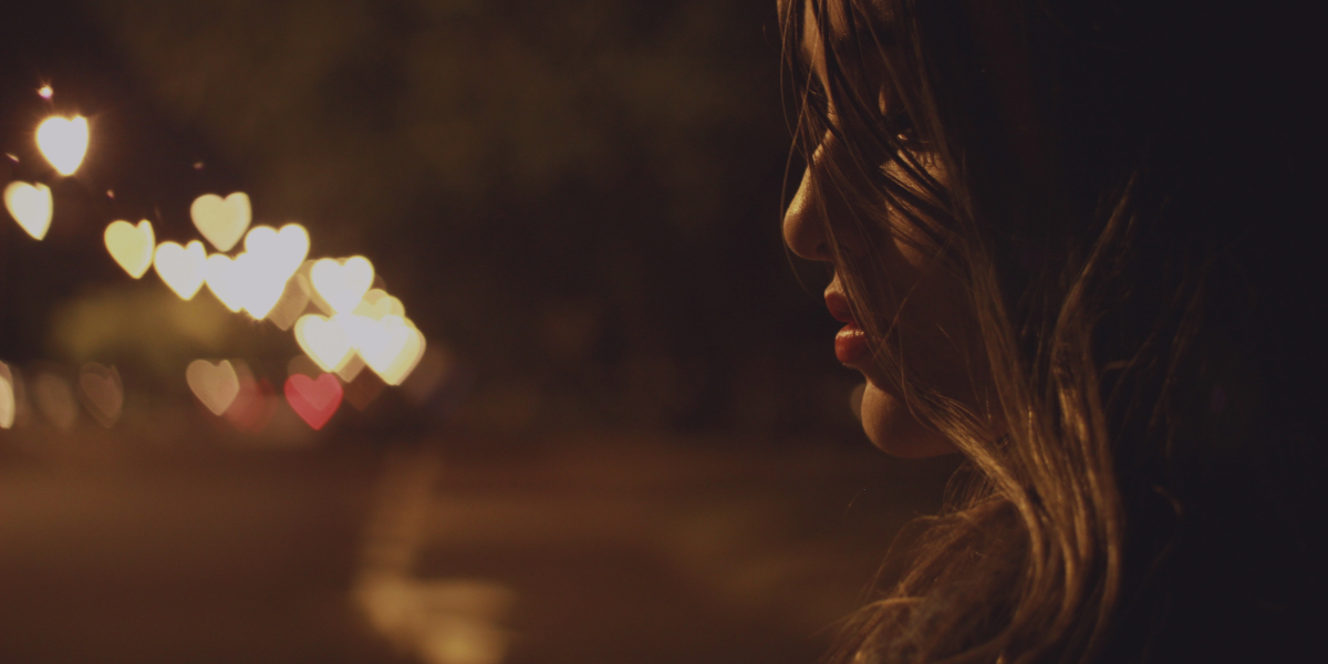 A woman standing in the dark. The sparkles of light in the distance have been distorted into heart shapes.