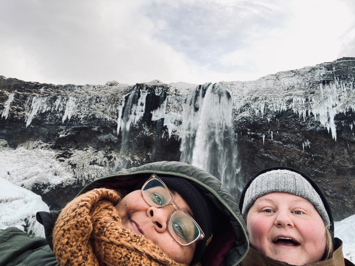 shea and Jane pose in front of a frozen waterfall. they're both wearing coats and knit caps. shea is wearing clear glasses and a scarf as well. They're both making kind of silly but also delighted faces!