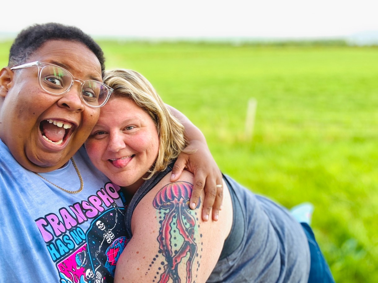 shea and their wife Jane are holding each other and smiling hugely on a field of bright green grass. shea is a Black nonbinary human with short hair and glasses. Jane is a white woman with a jellyfish tattoo on her arm and blonde shoulder length hair.