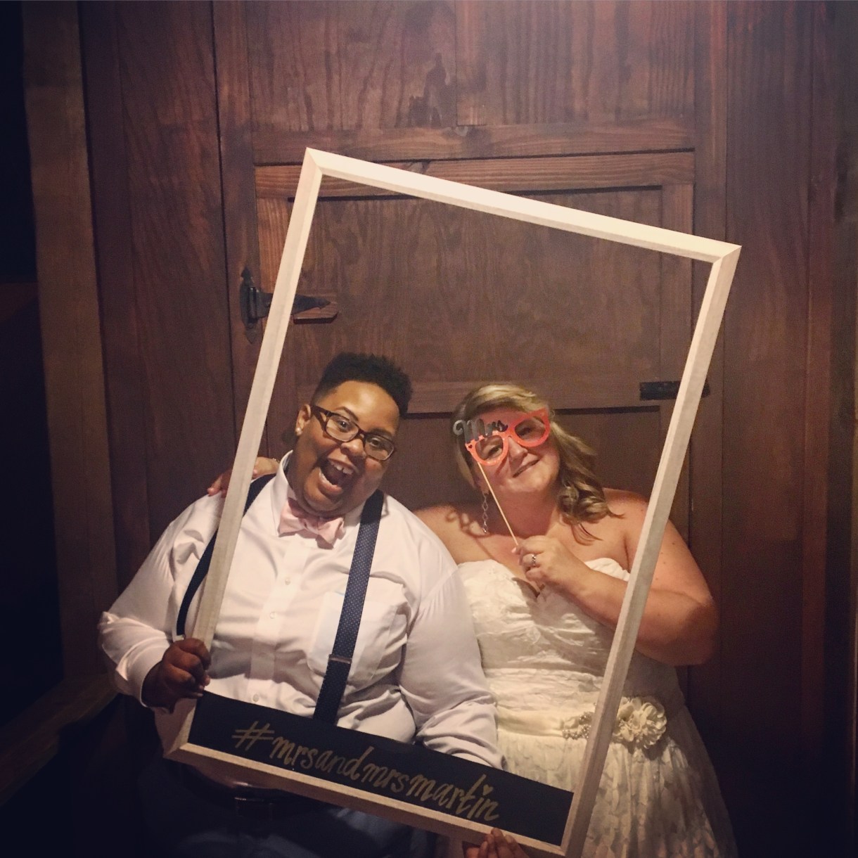 a wedding photo of shea and Jane. shea is wearing a white shirt, pink bowtie, suspenders, glasses, navy pants and a huge smile. Jane is wearing a gorgeous lacy wedding dress and is holding up glasses that are the type for photo props. shea is holding up a polaroid esque wooden frame with #mrsandmrsmartin written on its base. shea is a Black nonbinary human with short curly black hair and Jane is a white woman with shoulder length blonde hair that is curled and falling down to her shoulders
