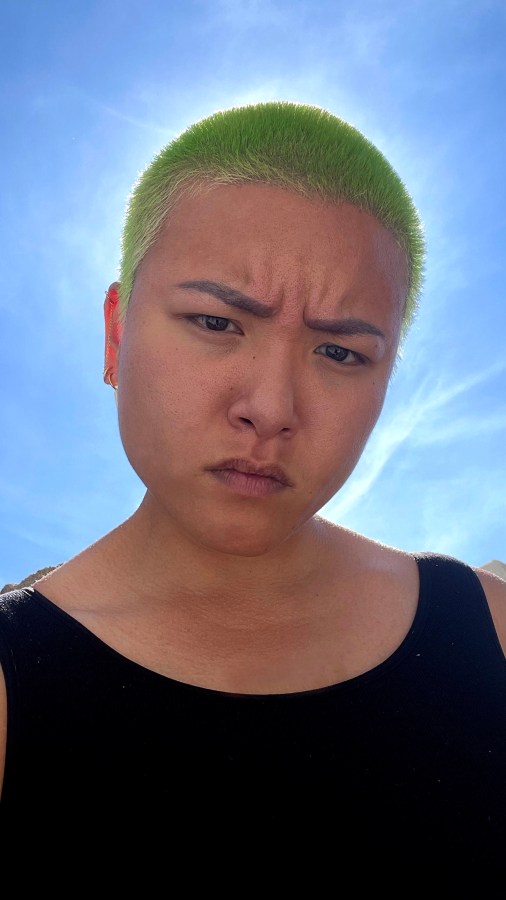 Viv, an Asian person, frowns at the camera with the sun behind them. They have green, buzzed hair and are wearing a black tank top.