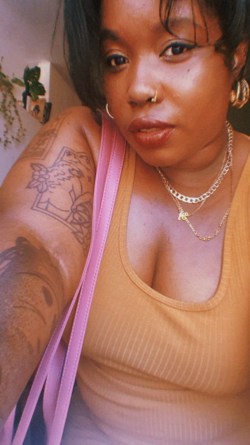 Shelli Nicole, a Black woman, poses for the camera, looking hot. She is wearing an orange tank and gold neckaces and hoop earrings and nose ring. She has a bag slung over her shoulder.