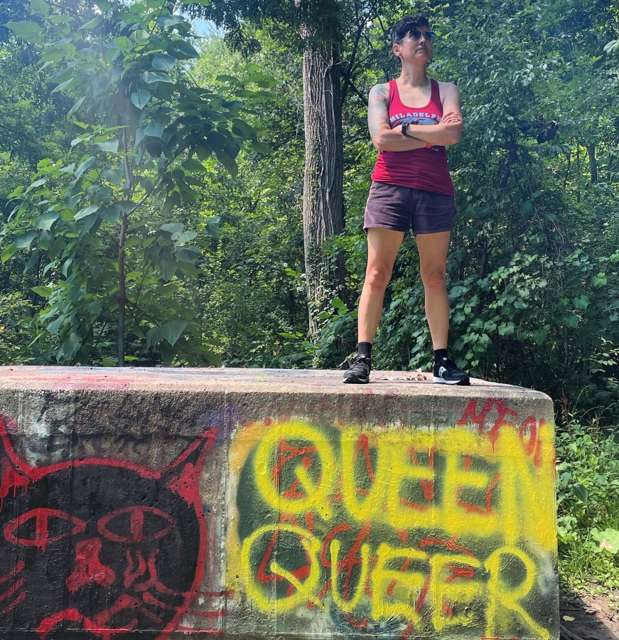 Tracy, an Asian woman, stands on top of a cement block graffittied with Queen Queer. She is wearing shorts, sneakers, and a tank. Her hair is short and dark and her arms are folded.