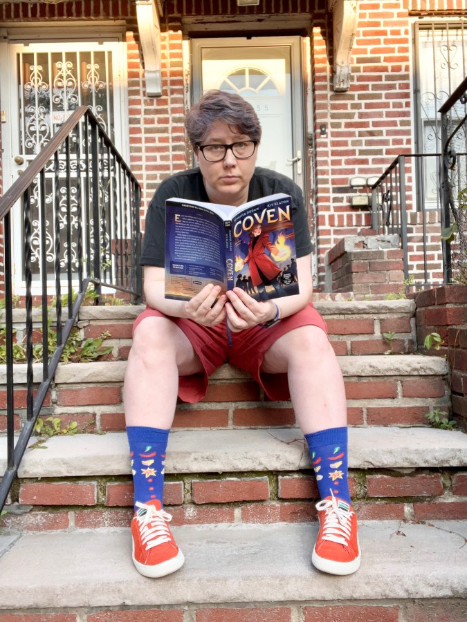 Heather is sitting on a stoop reading a book called Coven. She is a white woman with short gray hair, wearing a tee shirt, shorts, socks and sneakers.