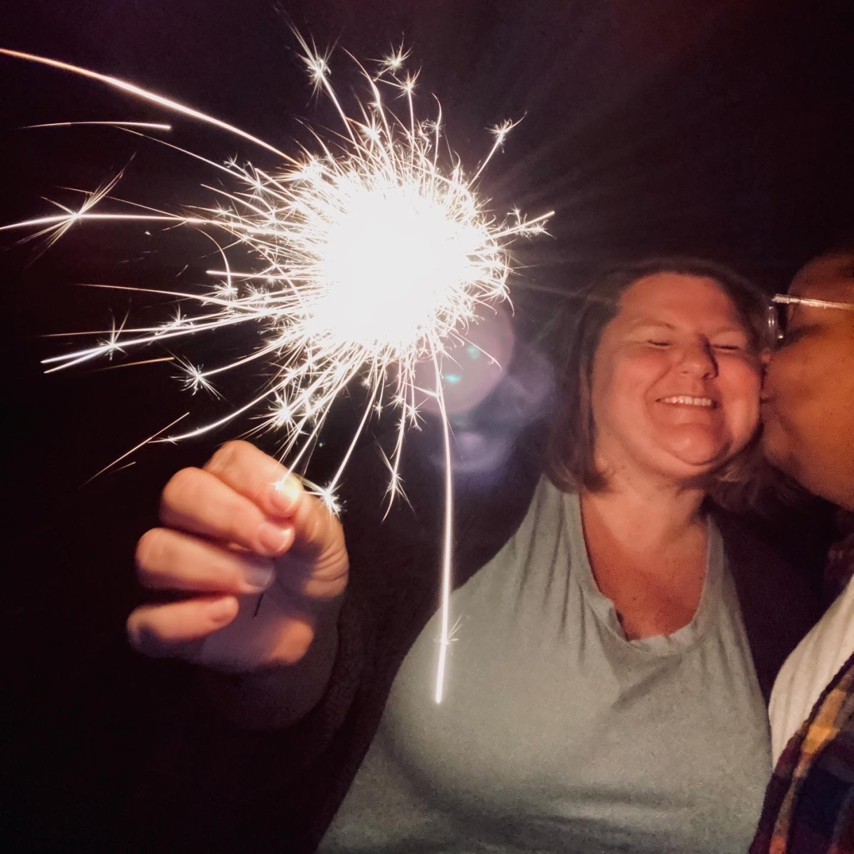 a photo with a central sparkler (like the fireworks kind). Jane is holding the sparkler toward the camera while shea kisses her on the cheek. the background is dark. it is night. they are both smiling.