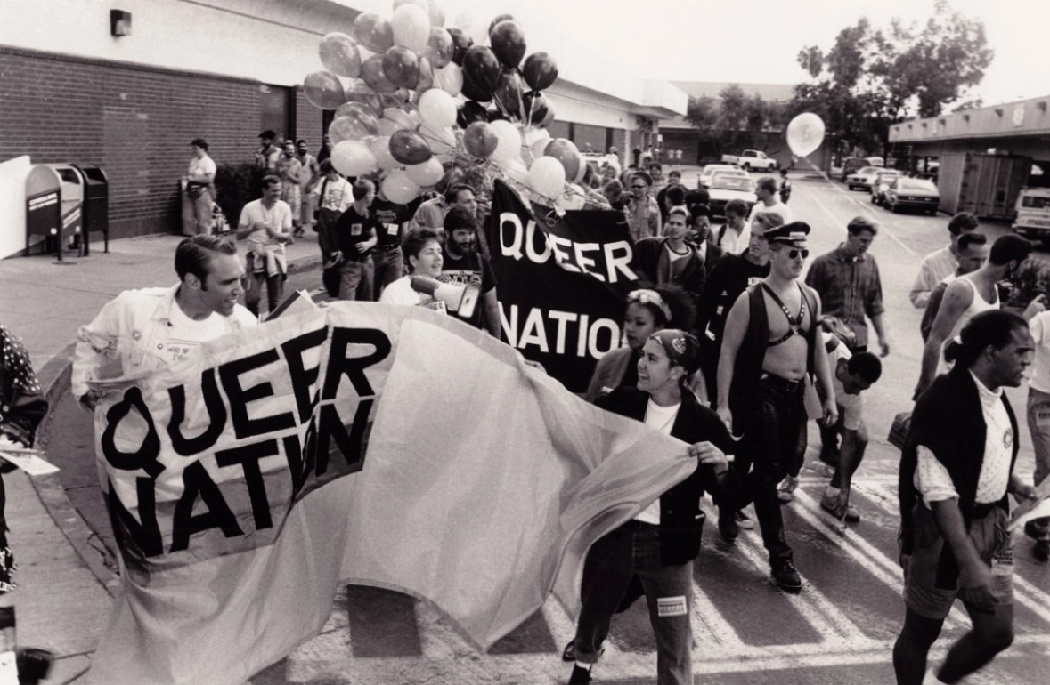 A black and white image shows a crowd of queer people in the street at a protest. Two people on the left hold a fabric sign that reads, "QUEER NATION" in black letters. Behind them, two more people hold a black fabric sign that reads, "QUEER NATION" in white letters. A large bundle of balloons is in the background. All of the participants are wearing typical street clothes, but one man is wearing a leather vest, a leather harness, a leather cap, leather pants and sunglasses. 