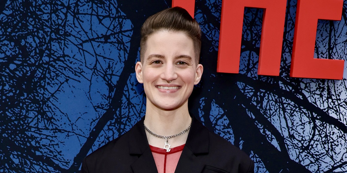 Theo Germaine, a white nonbinary trans person with short brown hair, is smiling on the They/Them red carped (there's a bright red They/Them sign behind them). They have a pink shirt, a metal choker, a black jacket, and a nose ring.