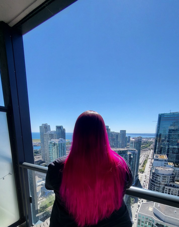 Valerie looks out over New York City as seen from behind on a balcony. Her hair is dyed bright pink. Valerie is a white woman.
