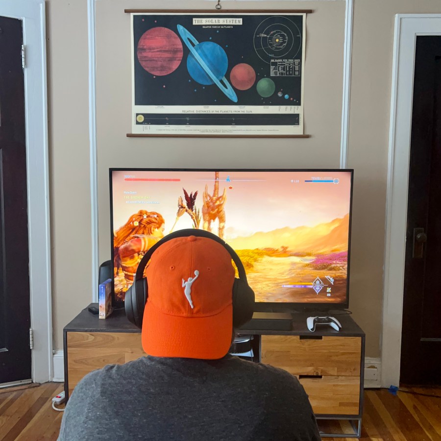 Heather plays video games, as seen from behind, wearing her WNBA baseball cap backwards. Heather is a white woman.