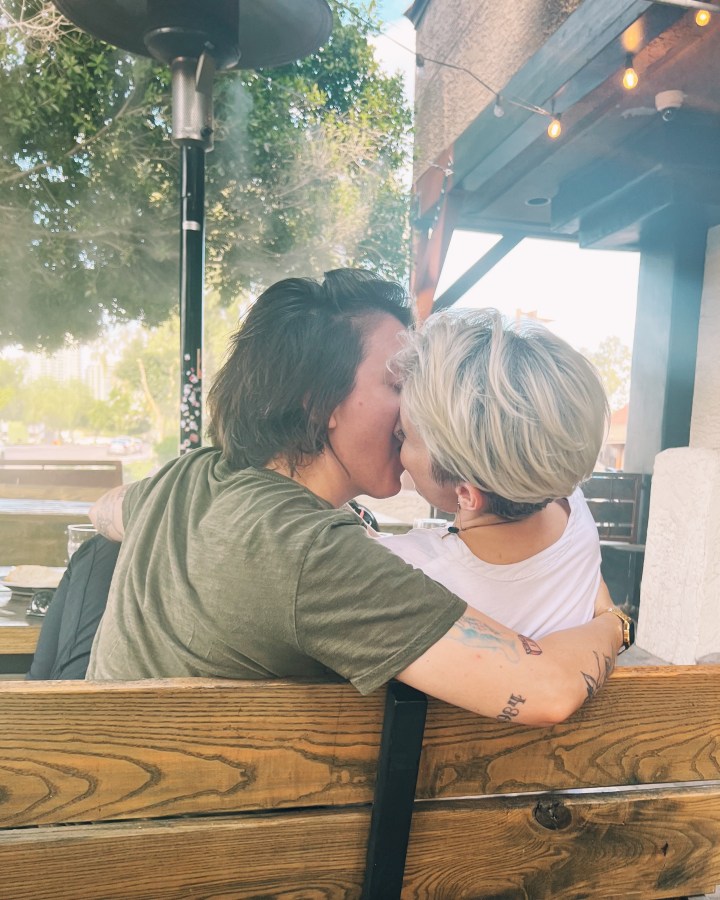 Laneia kisses her partner outside on a bench at some kind of establishment. Laneia is a white woman with short blonde hair wearing a white shirt.