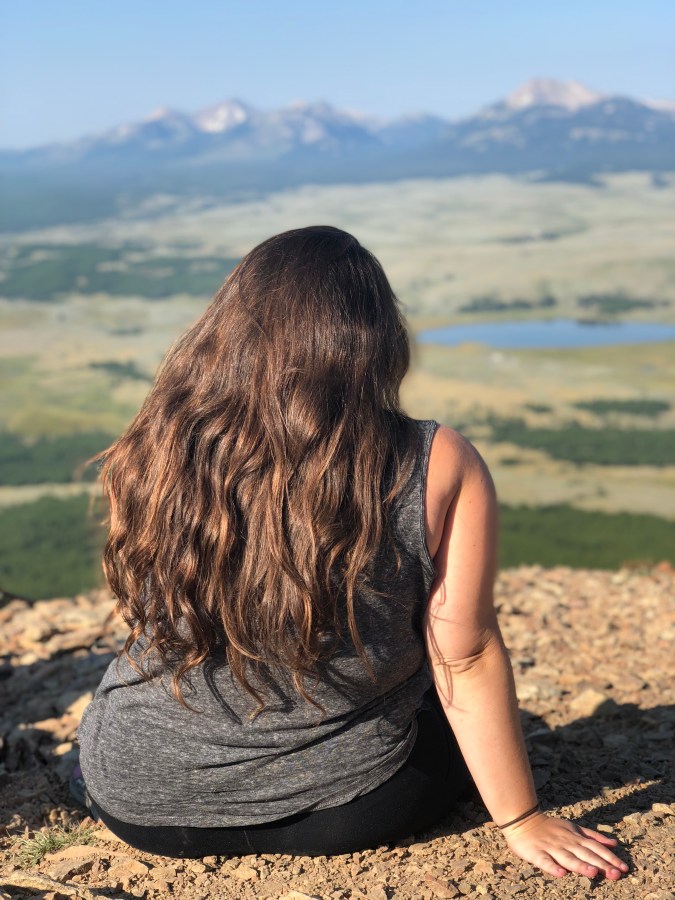Vanessa sits on a hill overlooking a beautiful natural landscape with mountains, a lake, and fields. She has long brown hair and is a white woman.