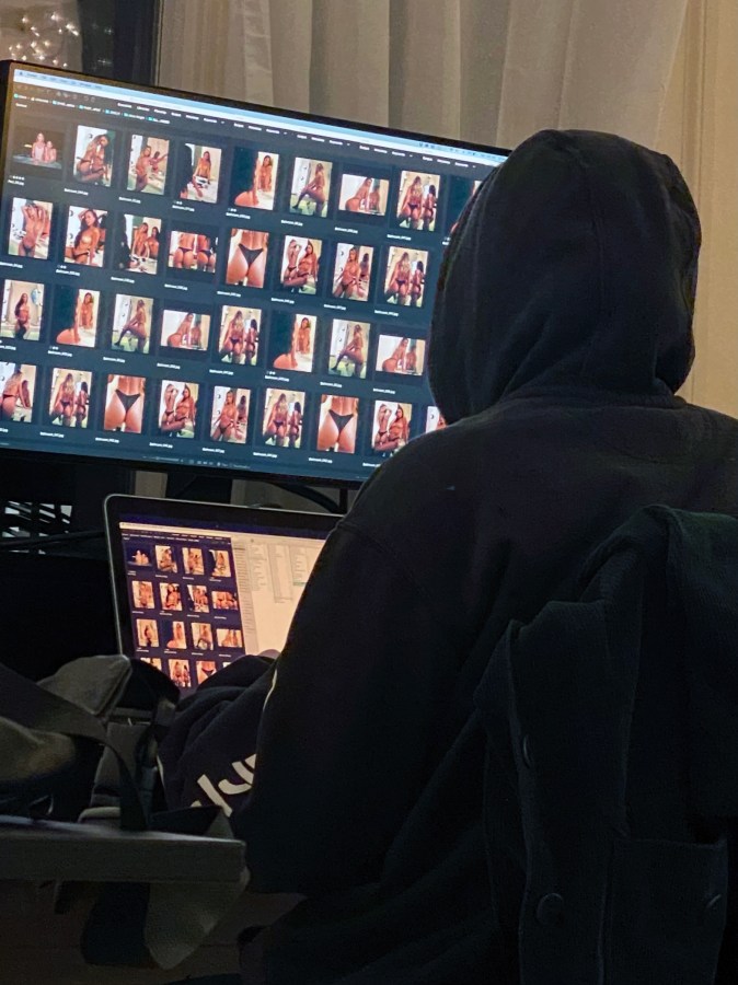 Viv is barely seen from behind here wearing a black hoodie, editing two computer screen fulls of model butts. Viv is an Asian nonbinary person.