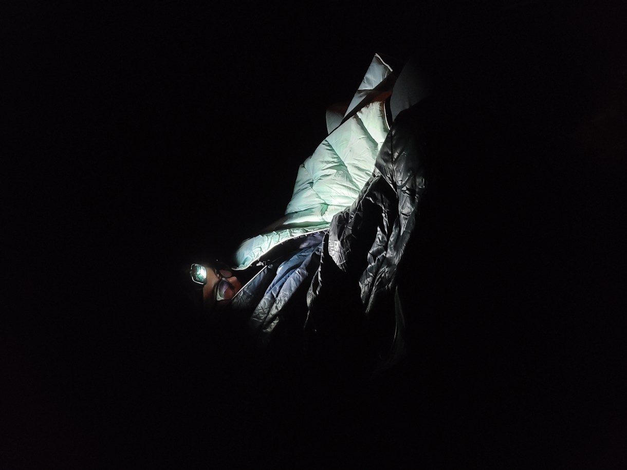 nico, a white genderqueer human, is bundled up in a blanket out of doors at night, illuminated only by the light of their headlamp