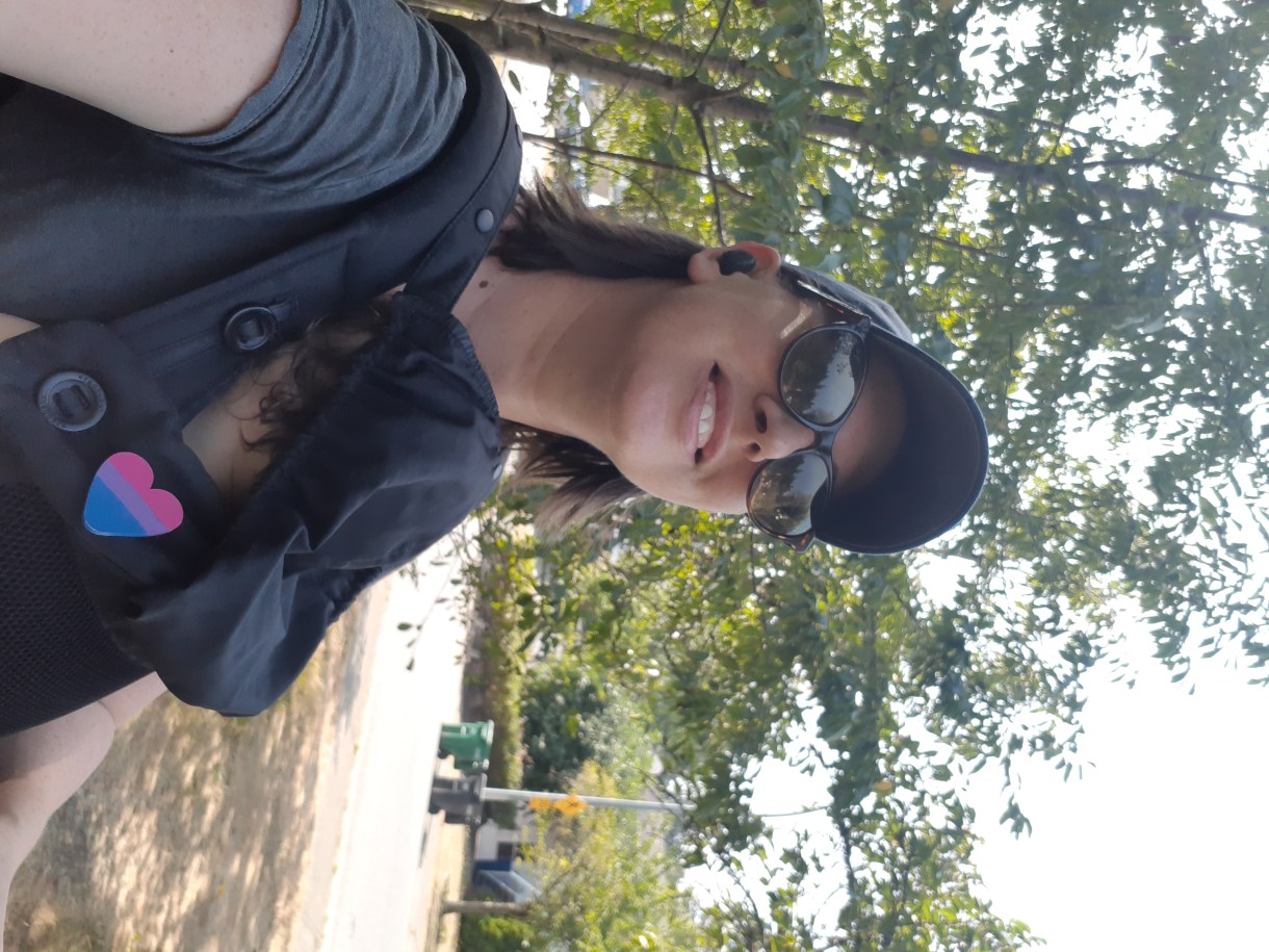 Casey, a white woman with brown hair wearing a baseball cap, walks through a park wearing heart-shaped sunglasses, with her baby in a carrier on her chest