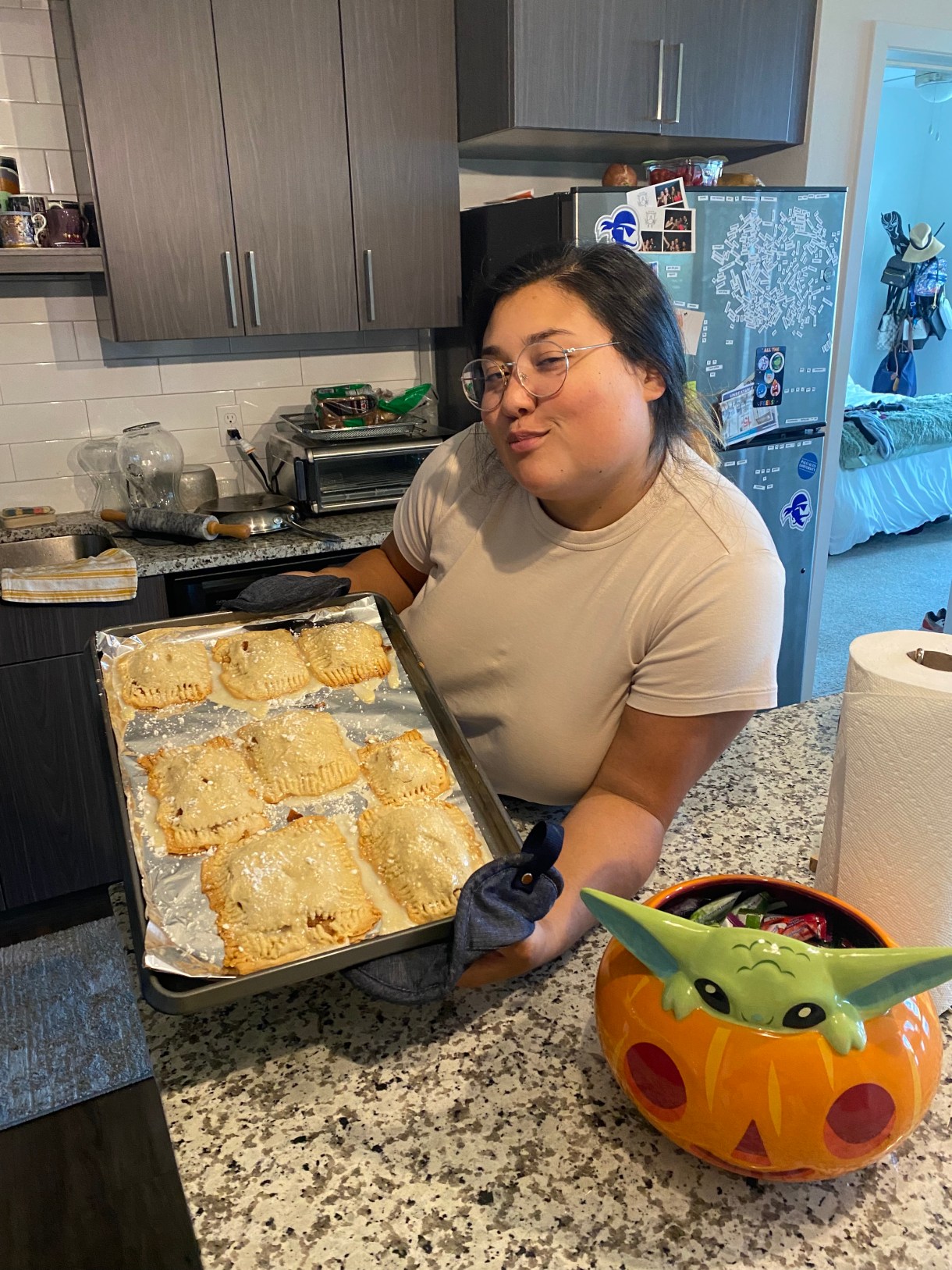 Em, a mixed race Asian human makes a pleased face while showing off some homemade pop tarts in a kitchen. they are wearing glasses and a taupe colored tee shirt.