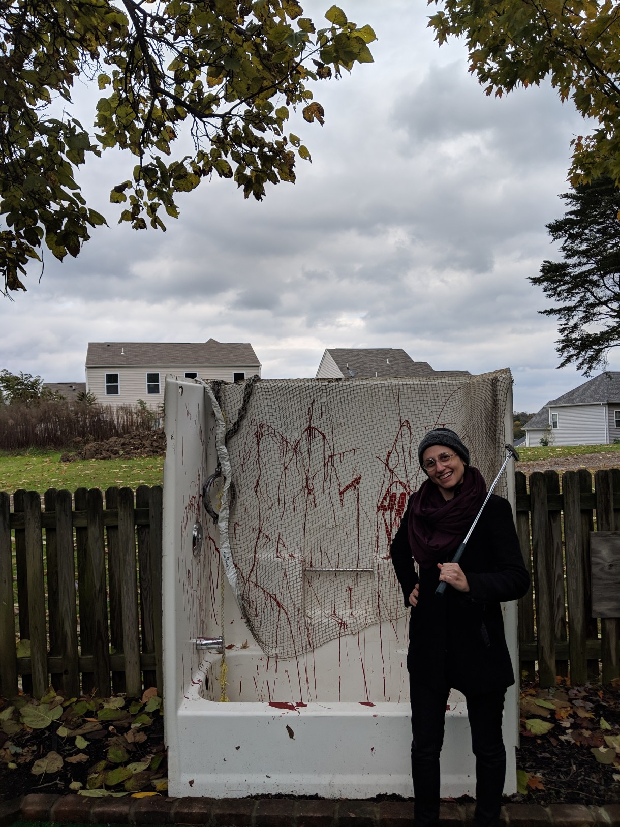 sadie, a white gender-nonconforming butch woman, grins happily in front of a shower placed eerily in front of a suburban fence on a cloudy fall day. she is wearing a beanie, scarf and black driving coat and has a putter over her shoulder. the shower and bath is covered in netting and fake blood