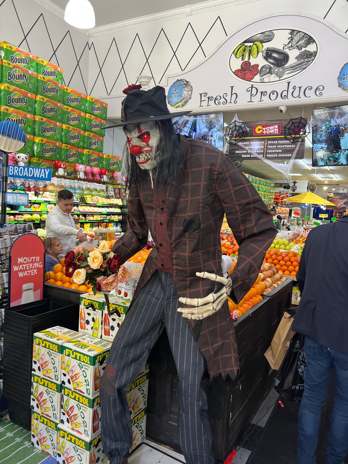a scary halloween clown skeleton with glowing red eyes and a plaid shirt hangs out in front of a fresh produce section at a grocery shop