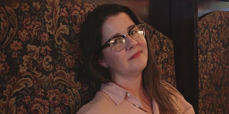 Kristen Arnett smiles while wearing glasses, propped against a brocade wall.