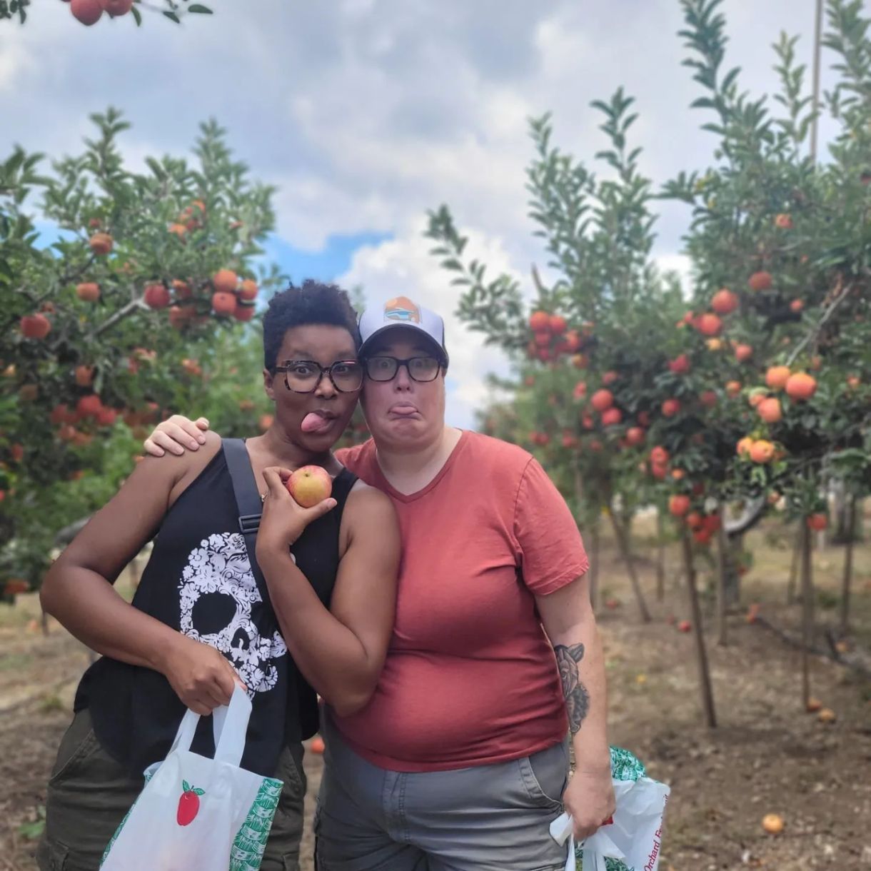 sai and her fiancee beth stand in an apple orchard and make funny faces while sai holds an apple. sa'iyda is a black woman with short hair and glasses wearing a skull tank top. beth is a white woman wearing a trucker hat, glasses and salmon tee shirt