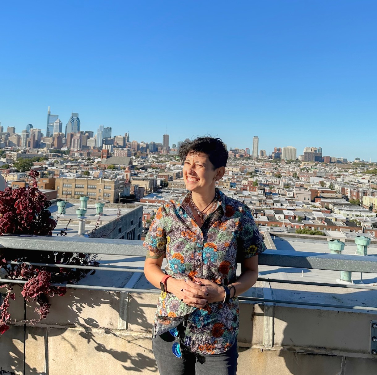 tracy stands, grinning, on a rooftop with Philadelphia in the back. she is an Asian woman wearing a colorful button up and orange wrist watch and jeans
