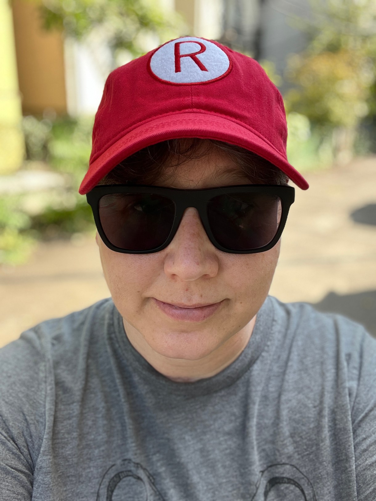 Heather takes a selfie outside in the sun, wearing a basebal cap with an R on it, sunglasses and a gray tee shirt. Heather is white and you cannot see her hair whichis all tucked under her hat in this photo