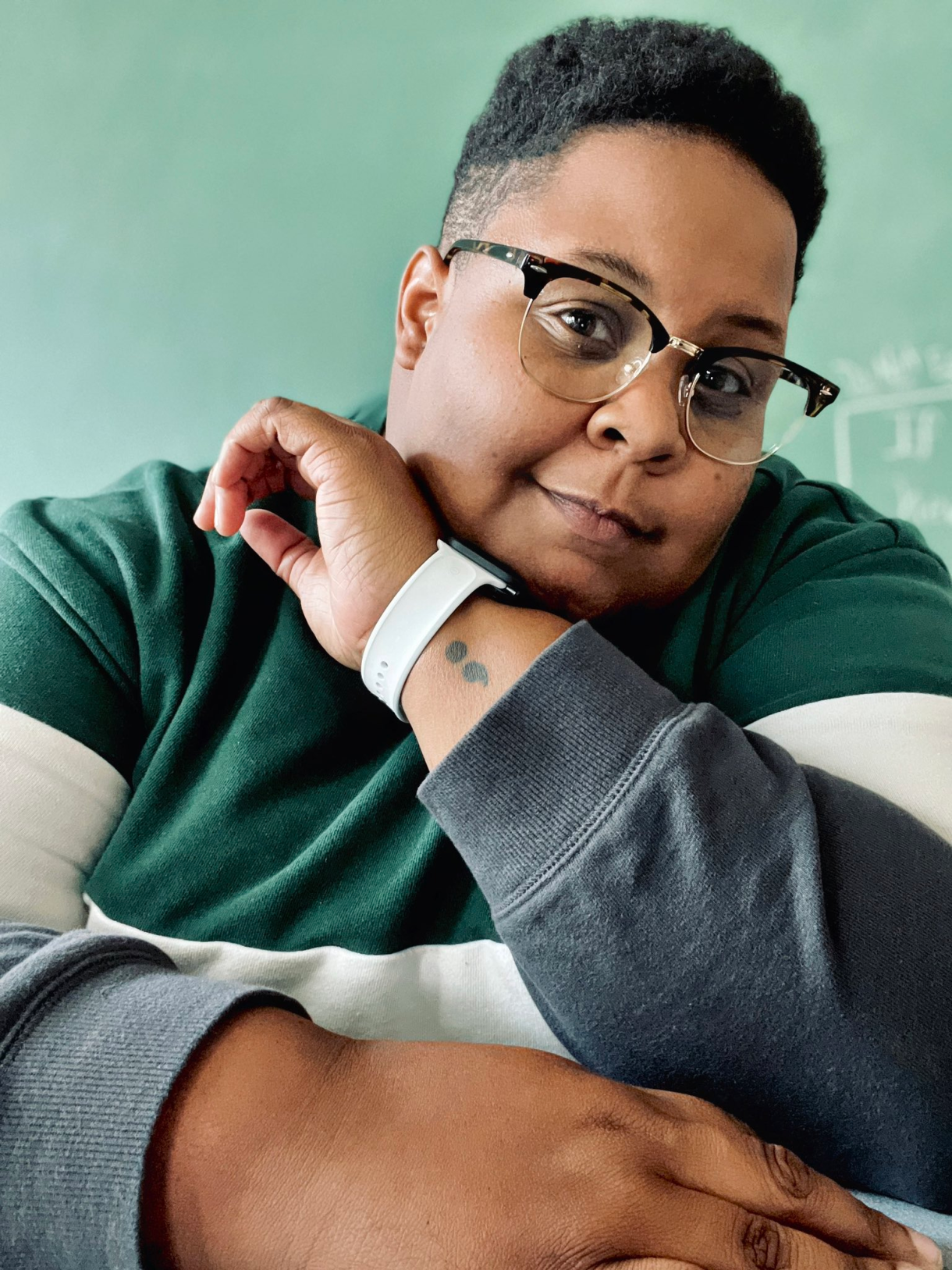 a photo of shea, a Black nonbinary human with short hair, shaved on the sides, toroise shell glasses, a white watch, a semicolon tattoo on their wrist, wearing a green and white striped polo. they are looking thoughtfully at the camera.
