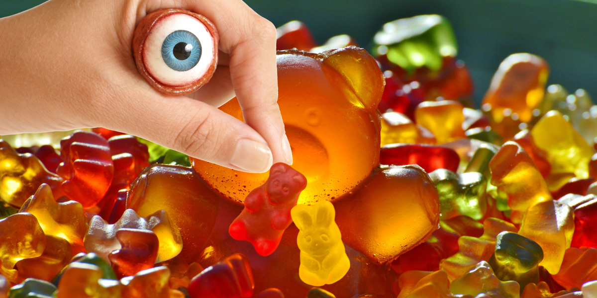 a collage with a background of gummy bears, including one big gummy bear, and a hand, with an eyeball creepily clutched in it, coming down to also pick up some gummies