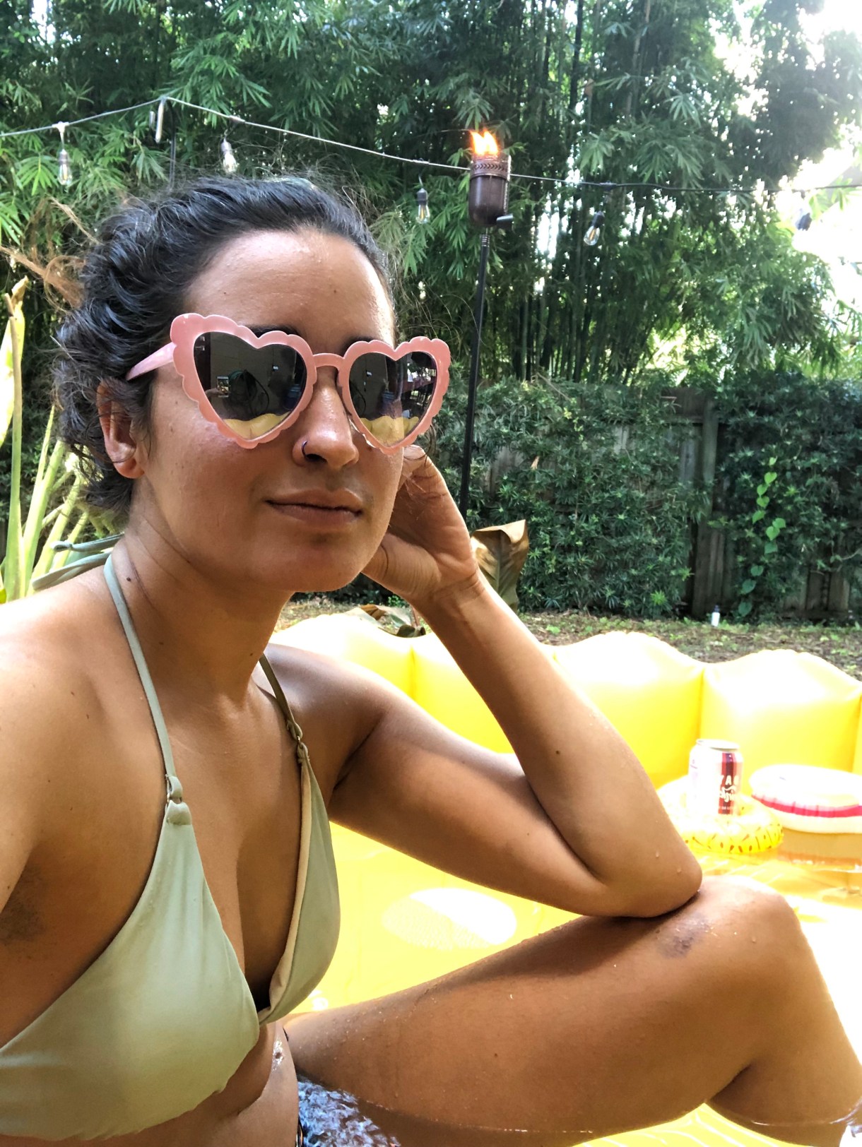 kayla, a south asian woman, wears a green bikini top and heart shaped sunglasses and sits in a kiddie pool in her backyard