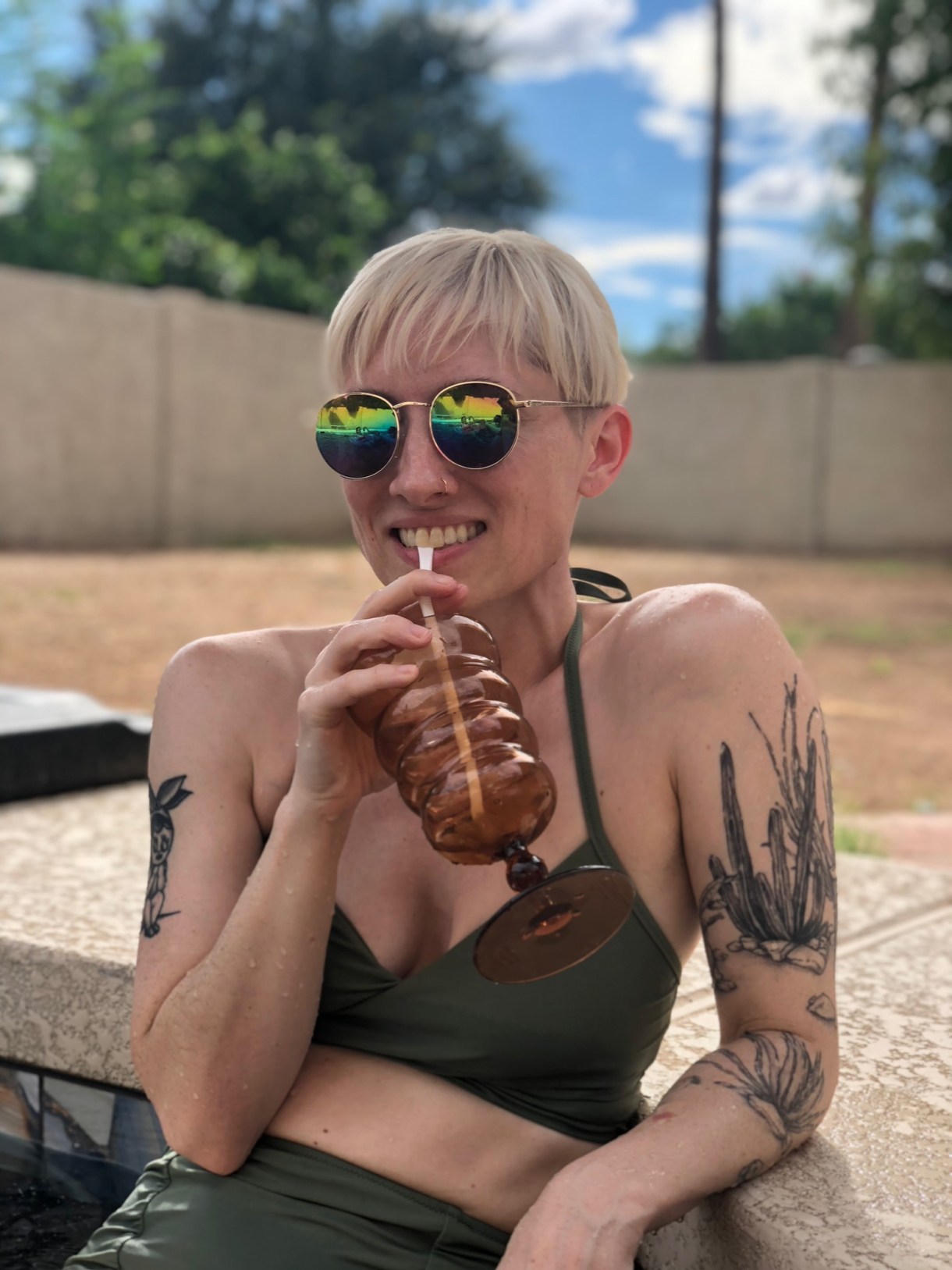 laneia, a white woman with blonde hair, wears mirrored sunglasses and smiles while siping a drink from a cup. she is wearing a grin bikini and leaning on the edge of a pool. you can see her tattoos
