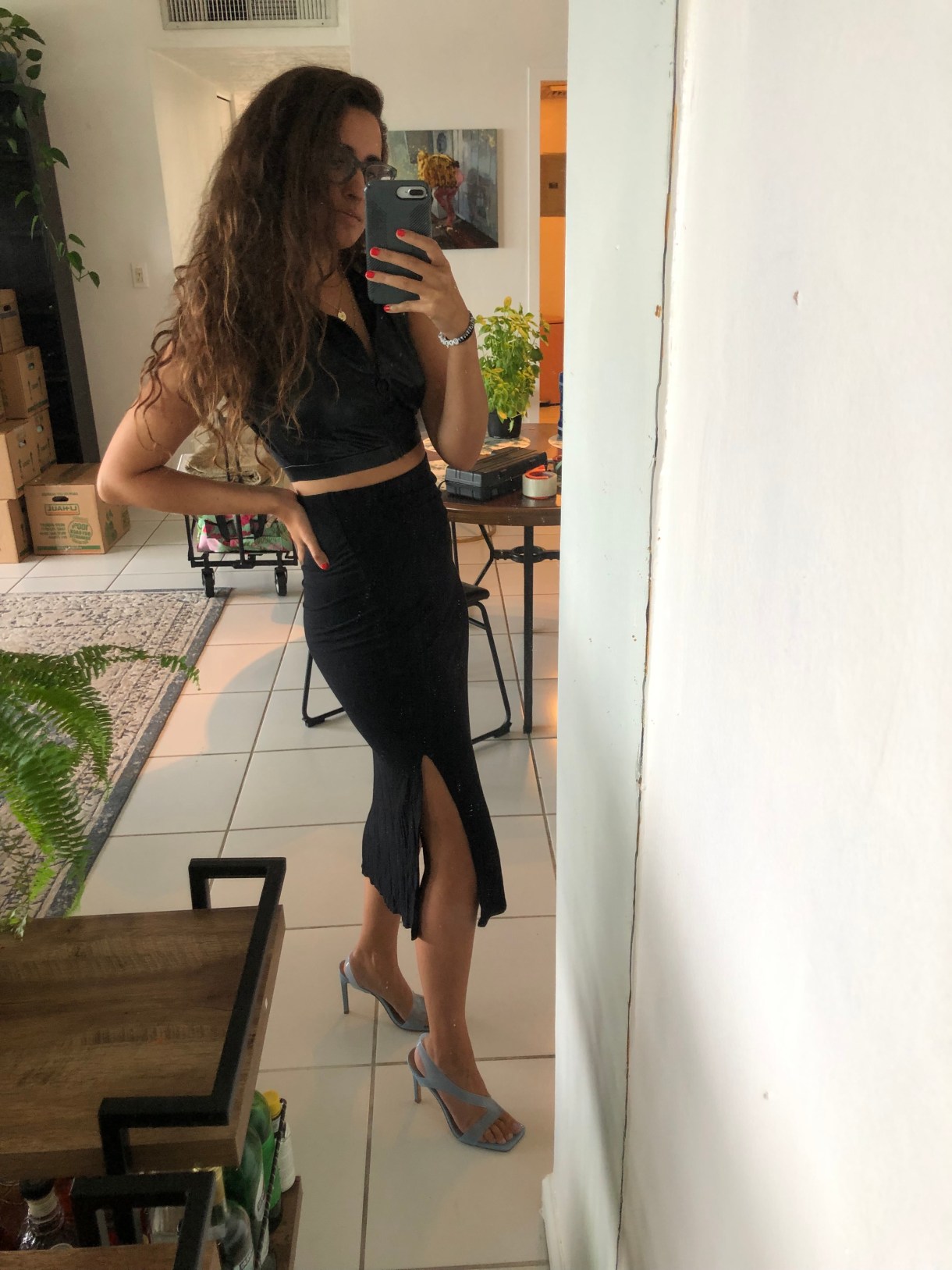 Kayla Kumari Upadhyaya wears a cropped pleather zip-up tank over a black slit midi jersey skirt and strappy pale blue square-toe heels. She has her hand on her hib and is taking a selfie in the mirror.